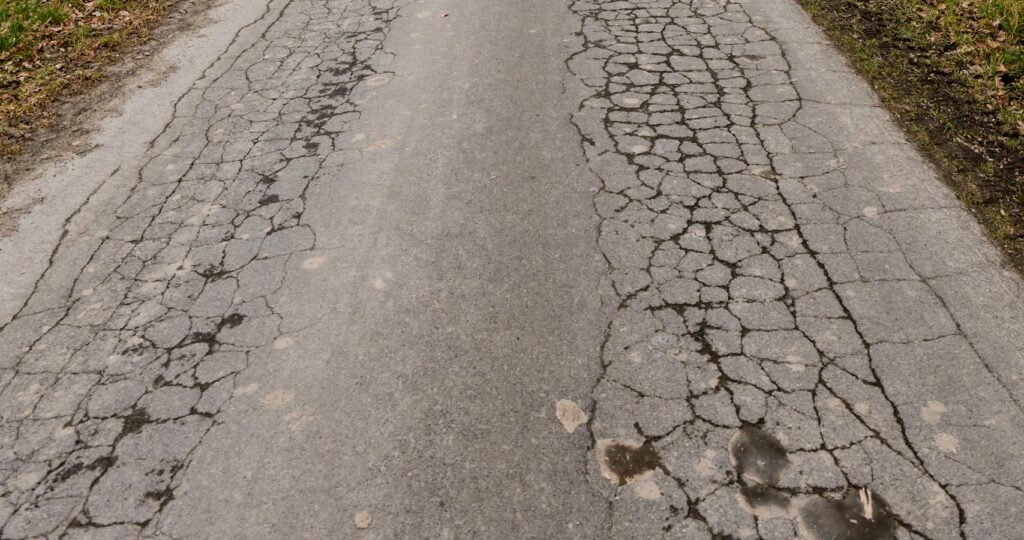 A cracked asphalt road leading through a barren winter landscape with trees on either side.