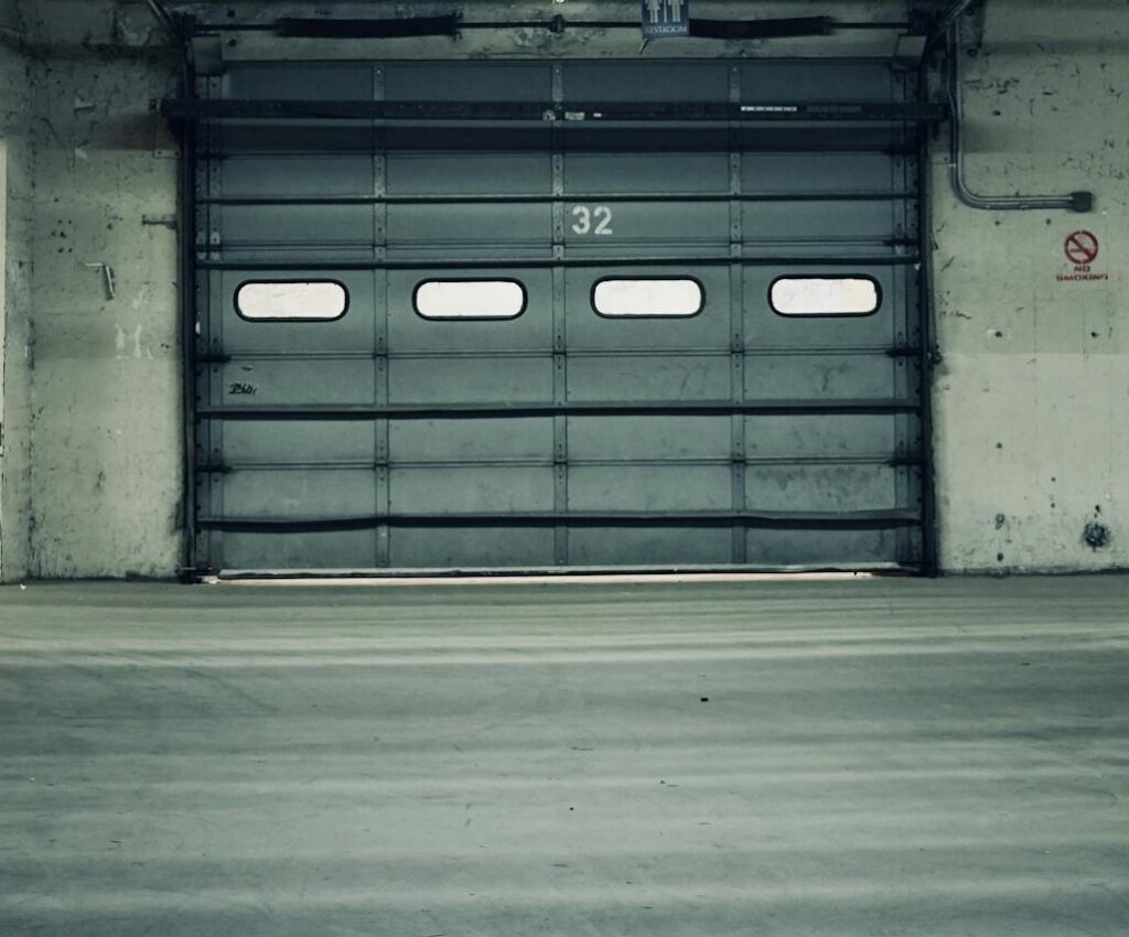 Empty industrial warehouse featuring stacked pallets and a closed garage door under fluorescent lighting.