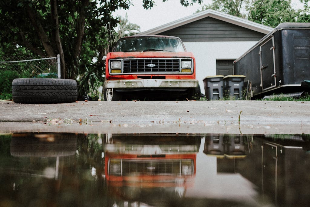 Old red truck with reflection on a wet driveway, showcasing rustic charm.